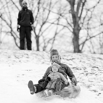 A man watching a woman and little kid sledding down a snowy hill.