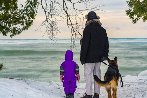 A person with a child and a dog in the snow at the lakefront.