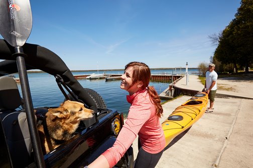 A couple carrying a kayak next to a Jeep with a dog in it.